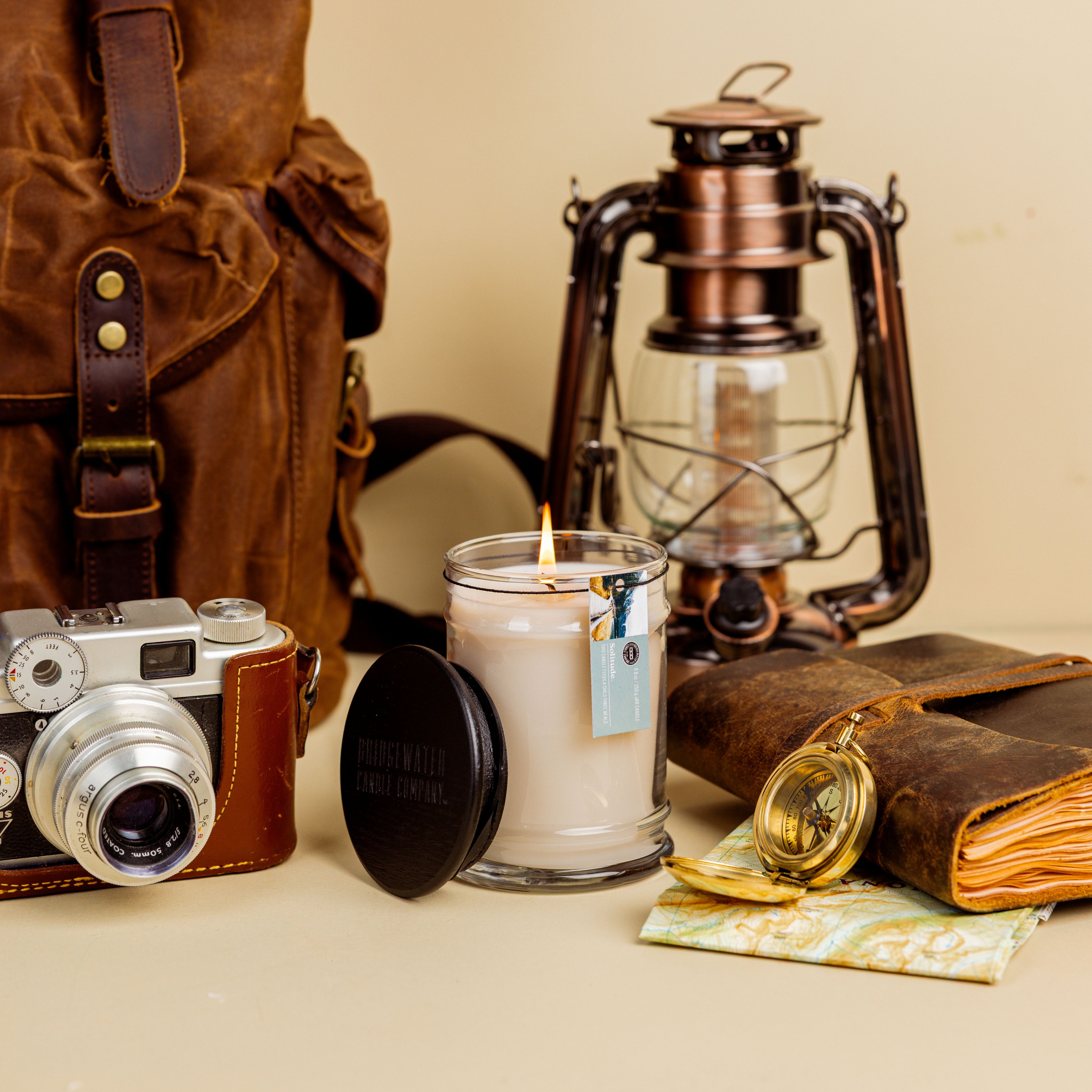 A still life of travel items includes a vintage camera, a brown backpack, a lit Solitude Small Jar Candle by Bridgewater Candle made with soy-blend wax, a lantern, leather-bound journal, map, and brass compass on a beige surface.