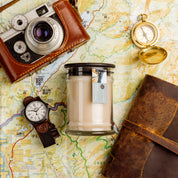 A vintage camera, watch, leather-bound journal, brass compass, and a Solitude Small Jar Candle by Bridgewater Candle are arranged on a colorful map, evoking a sense of travel and adventure.