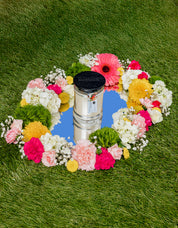 A white urn with a black lid sits on a round mirror amid colorful flowers. Nearby, the Bridgewater Candle High Bloom 8oz Small Jar Candle with soy-blend wax gently scents the green grass setting.