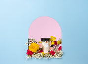 The Bridgewater Candle High Bloom 8oz Small Jar Candle, made with soy-blend wax in a glass jar with a black lid, sits amid colorful flowers in a small arched alcove with a blue background and pink interior.