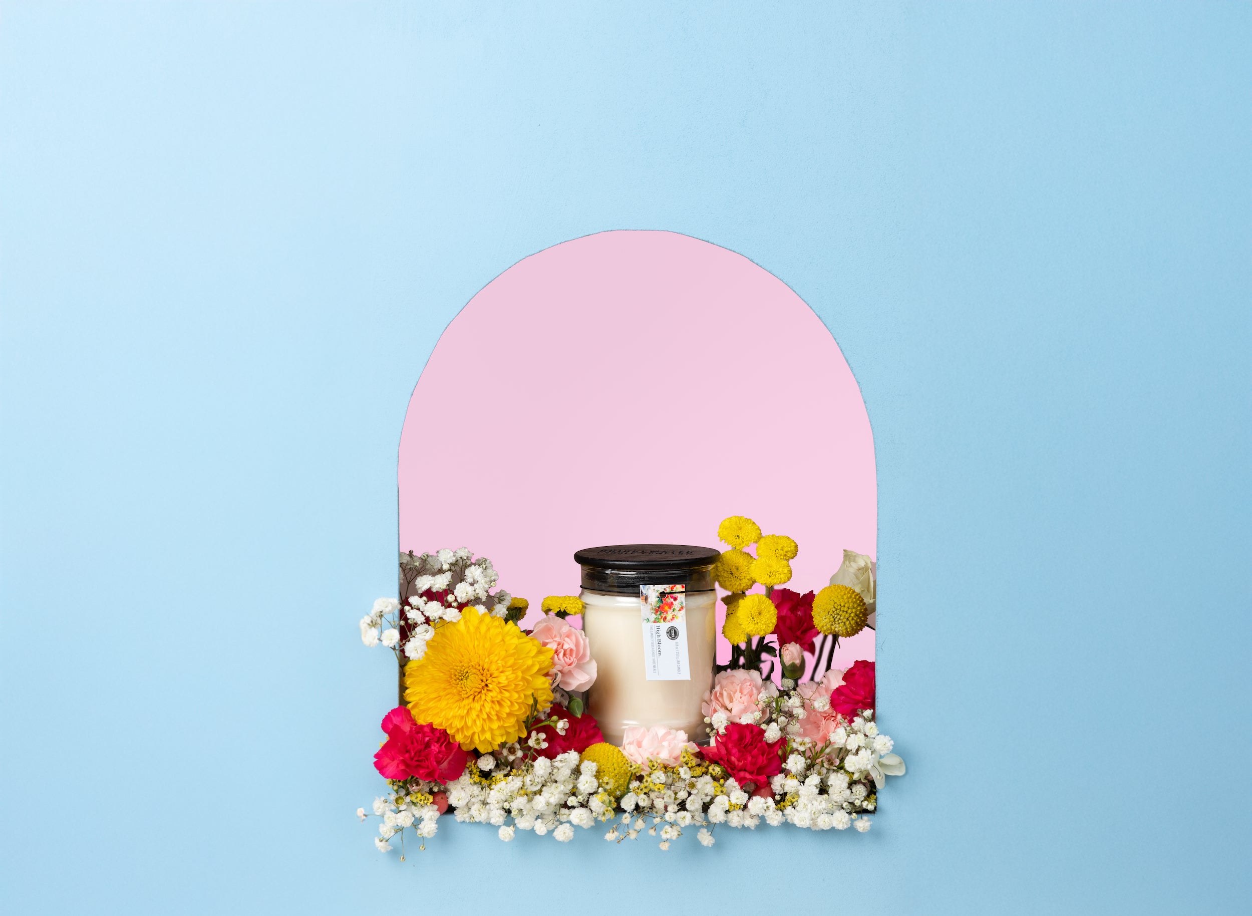 The Bridgewater Candle High Bloom 8oz Small Jar Candle, made with soy-blend wax in a glass jar with a black lid, sits amid colorful flowers in a small arched alcove with a blue background and pink interior.