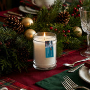The Bridgewater Candle "I’ll Be Home" 18oz Large Jar Candle, made from soy-blend wax, glows in a glass jar on a festive table with evergreens, pinecones, gold ornaments, and red berries, adding cozy fragrance to your holiday setting.