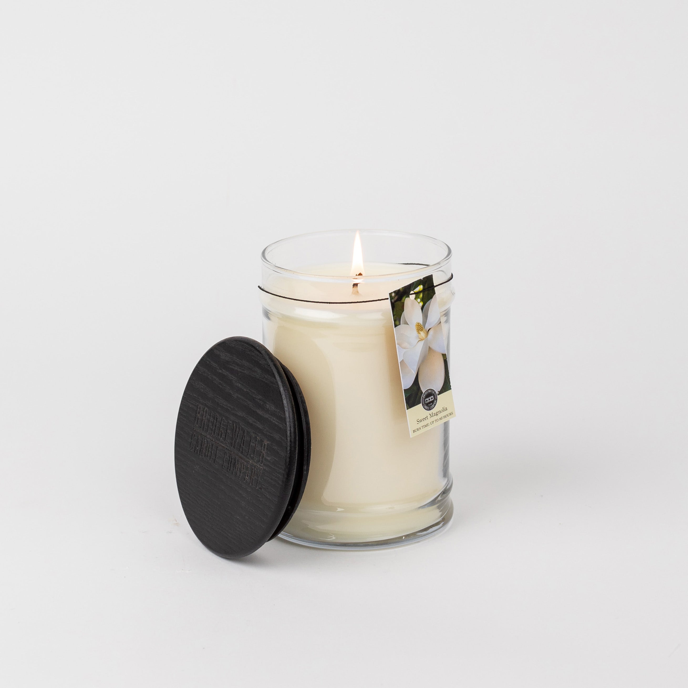 A Sweet Magnolia 18oz Large Jar Candle by Bridgewater Candle Company sits lit in a clear glass jar with a black wooden lid beside it. A tag with white flowers suggests the Sweet Magnolia scent, all set against a plain white background.