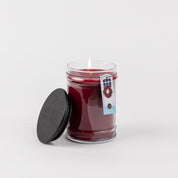 A lit Bridgewater Candle Company Welcome Home 18oz Large Jar Candle in a glass jar with a black lid beside it. The label shows a window with a wreath, and the candle is set against a plain white background.