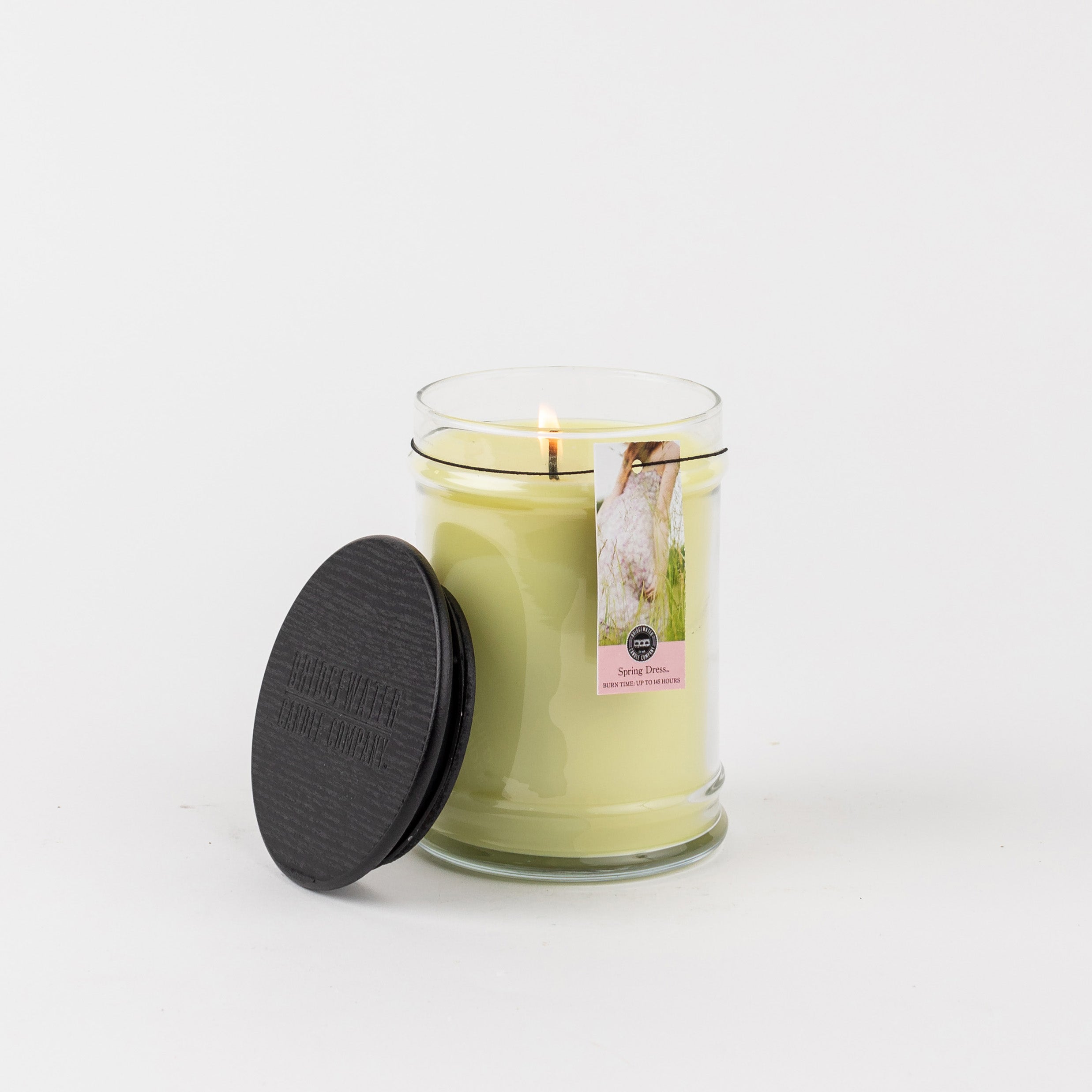 A lit Spring Dress 18oz Large Jar Candle by Bridgewater Candle Company sits in a clear glass jar with a black lid nearby. The floral label stands out against the plain white background, highlighting this elegant fragrance candle.