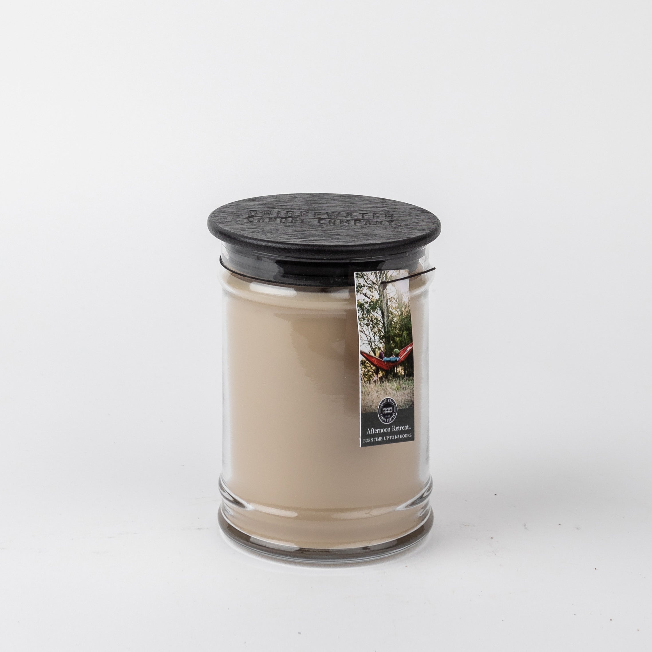 The Bridgewater Candle Company Afternoon Retreat 18oz Large Jar Candle features a beige fragrance in a clear glass jar with black lid and label with a person in a hammock. Enjoy its calming scent in any room. Background is plain white.