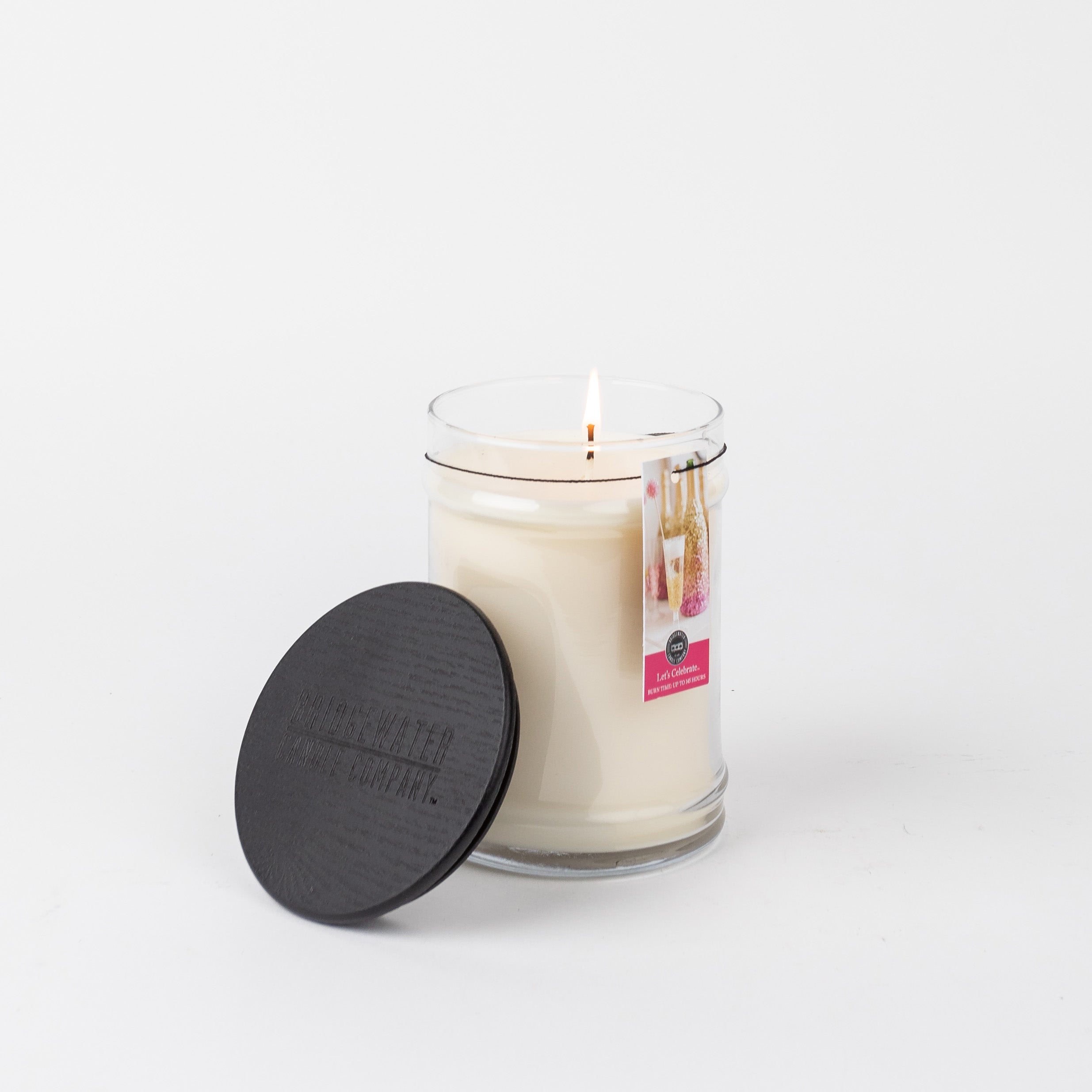 The Bridgewater Candle Company Let's Celebrate 18oz Large Jar Candle burns in a clear glass jar with a black lid and tag, set against a plain white background—ideal for enjoying your favorite fragrance.