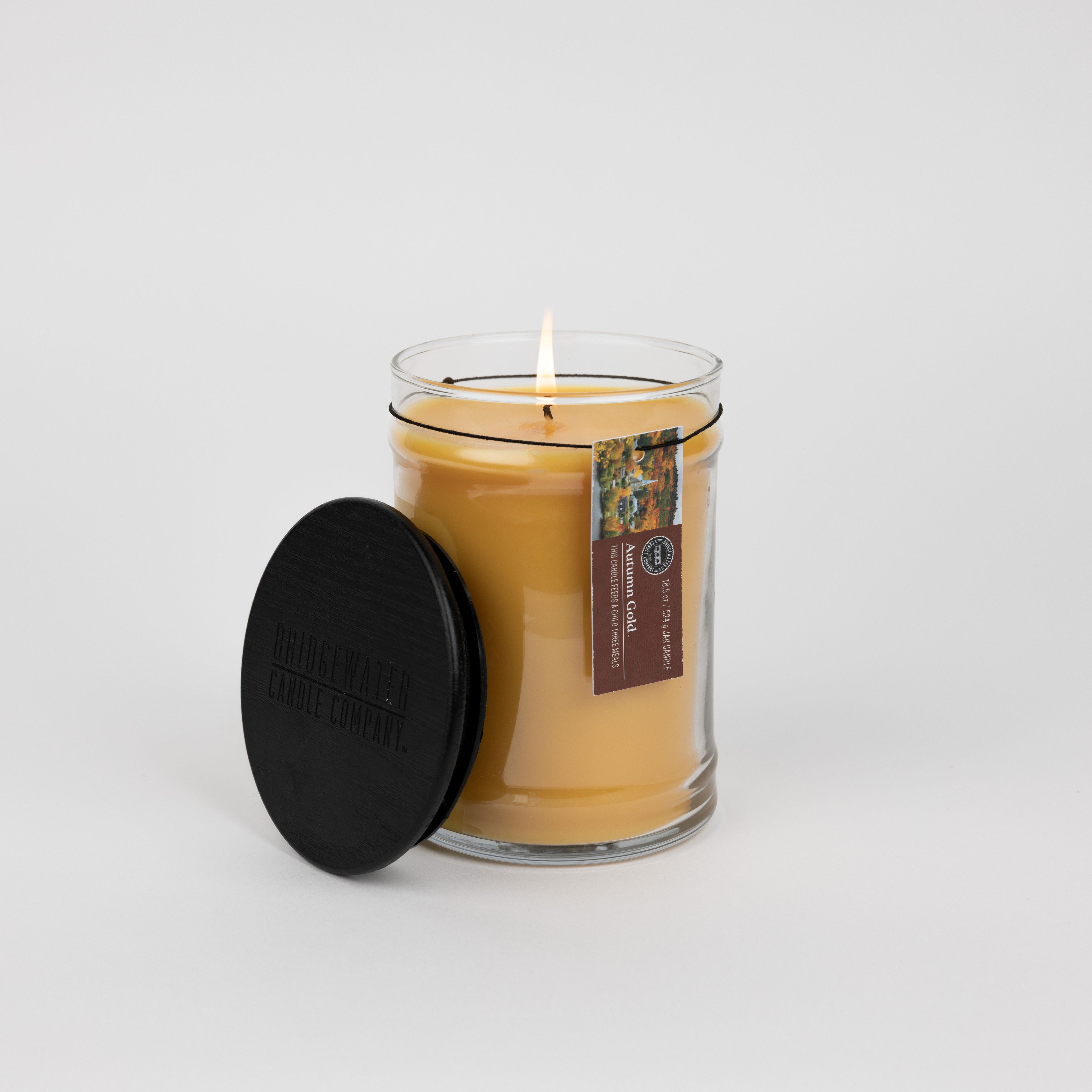 The Autumn Gold Large Jar Candle by Bridgewater Candle, made with soy-blend wax, glows in a clear glass jar with a black lid propped beside it. A decorative label accents the jar, set against a plain white background.