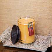 The Autumn Gold Large Jar Candle by Bridgewater Candle, made with soy-blend wax in a clear glass jar with a dark lid and hanging tag, leans on a patterned tray against a beige textured background.
