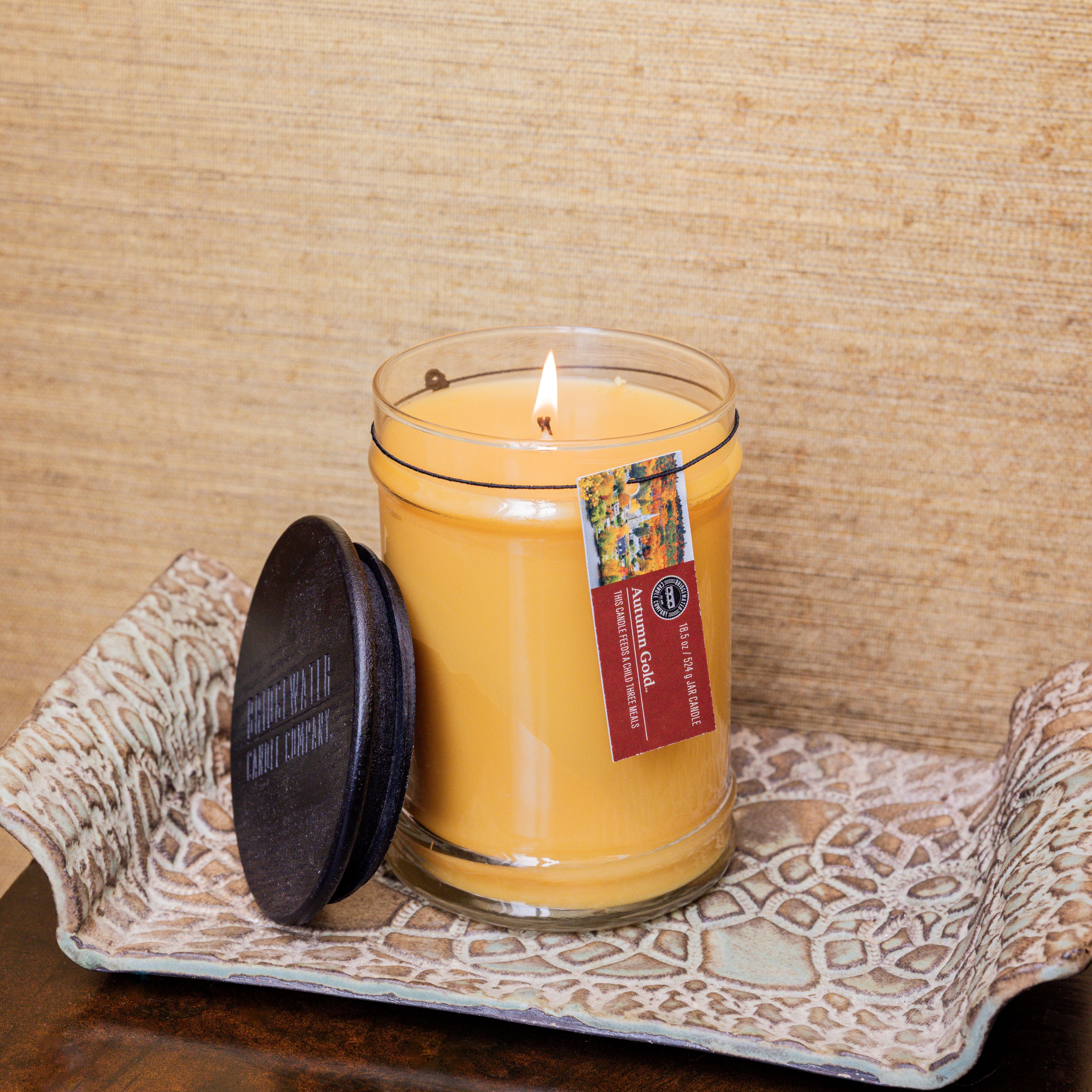 The Autumn Gold Large Jar Candle by Bridgewater Candle, made with soy-blend wax in a clear glass jar with a dark lid and hanging tag, leans on a patterned tray against a beige textured background.