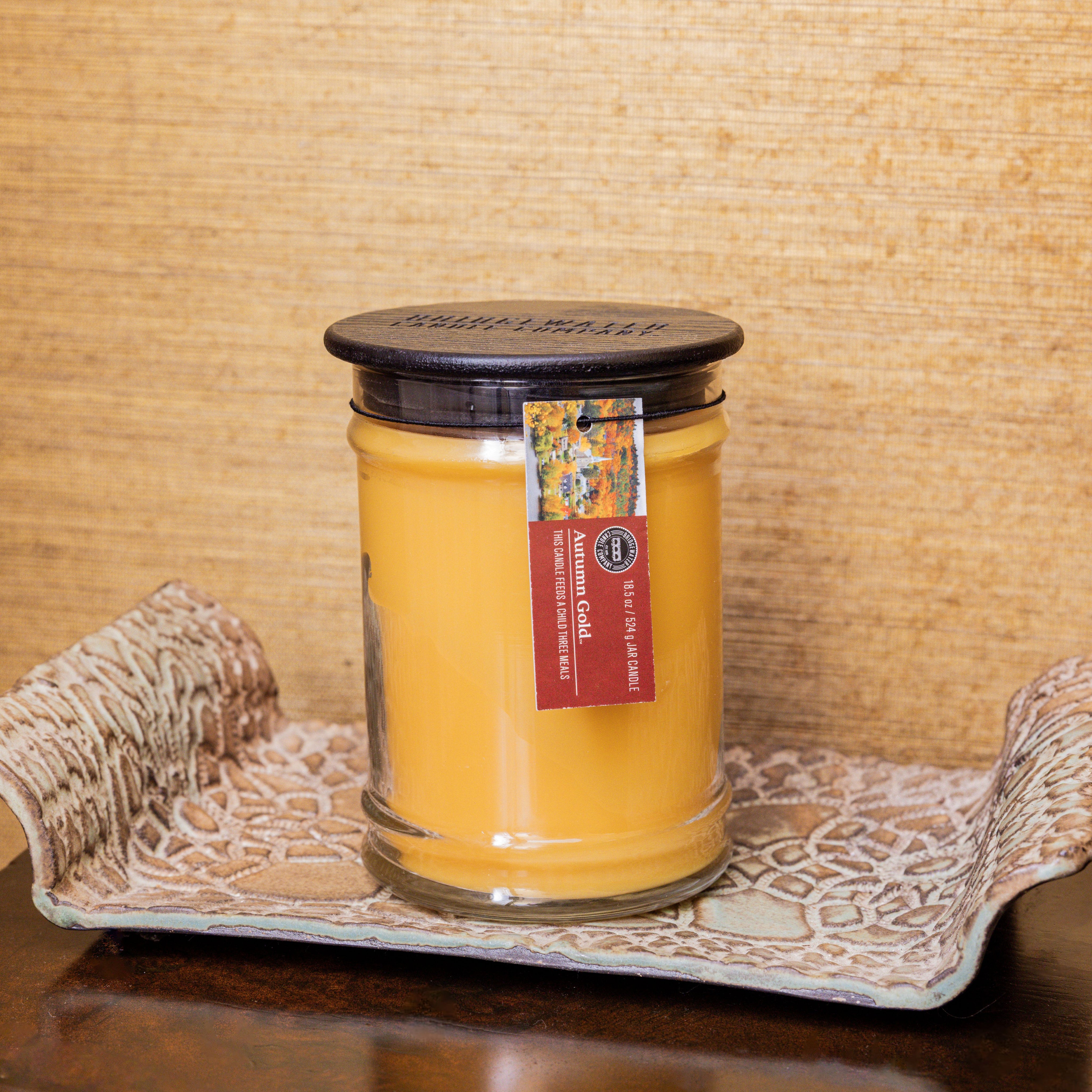 The Bridgewater Candle Autumn Gold Large Jar Candle, featuring orange soy-blend wax and a black lid, rests on a decorative tray. Its label displays Autumn Gold, set against a textured tan wall in the background.