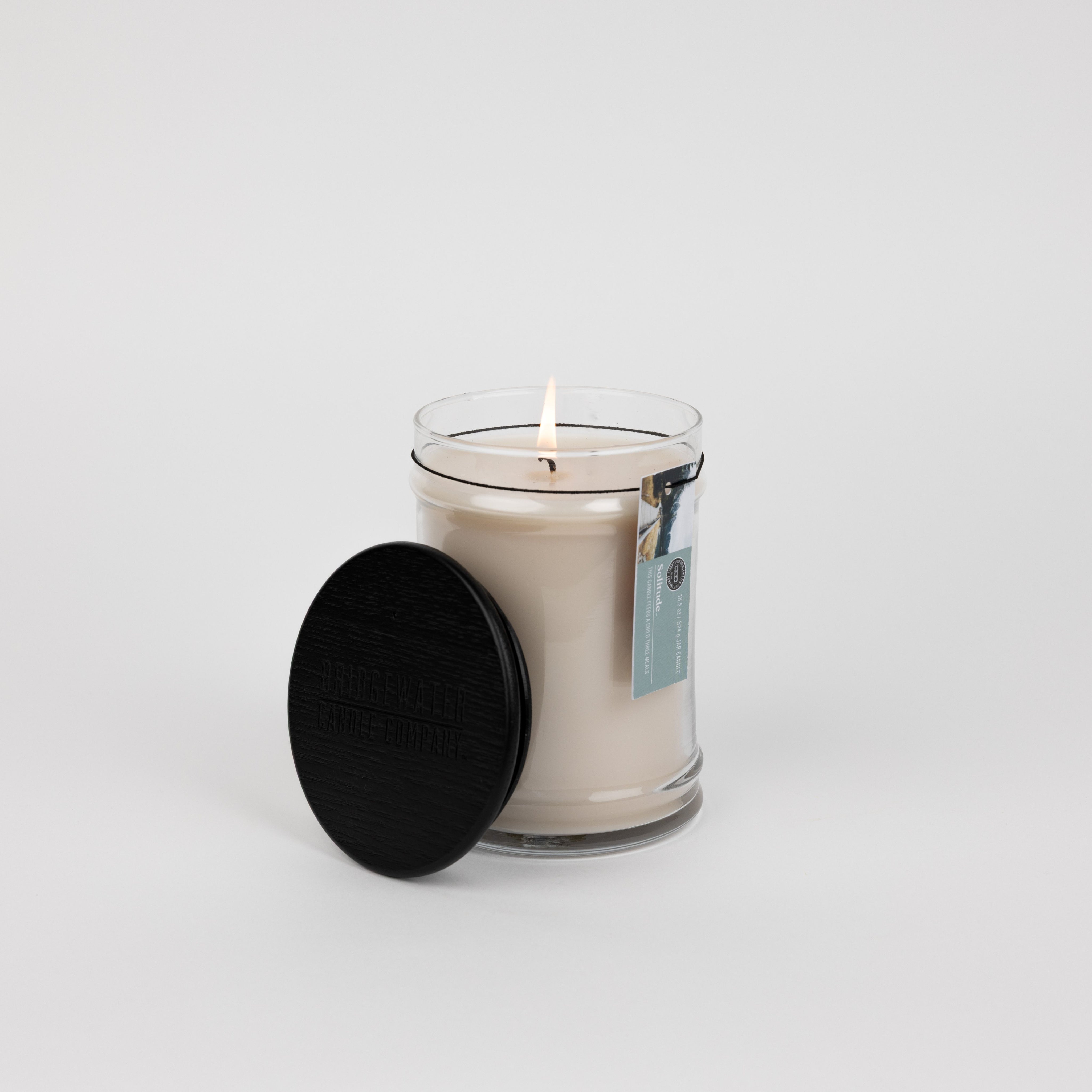 A lit Bridgewater Candle Solitude Large Jar Candle with white soy-blend wax glows in a clear glass jar, topped by a black lid resting on its side. A tag hangs from the jar, all set against a plain white background.