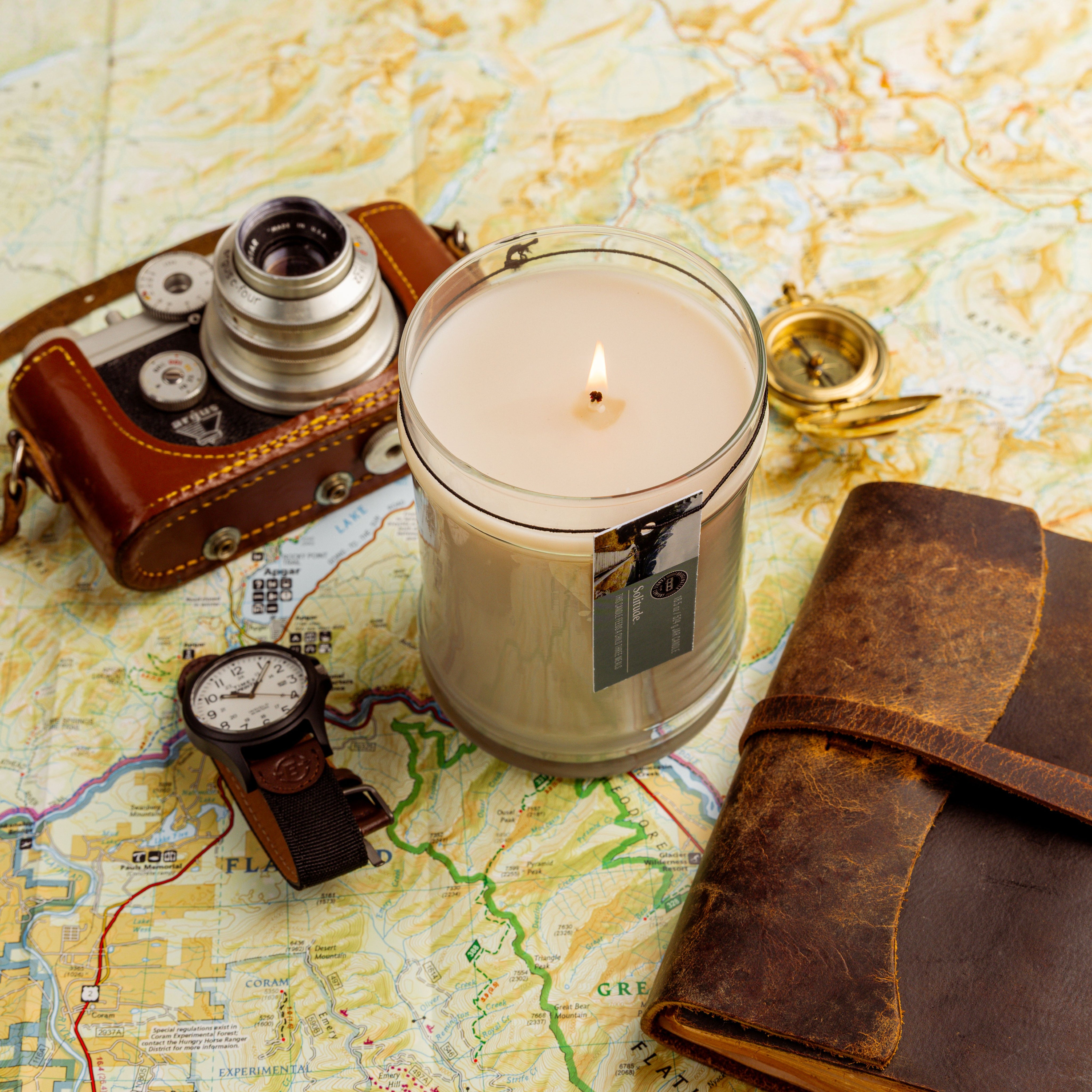 The Solitude Large Jar Candle by Bridgewater Candle, made with soy-blend wax, glows in a glass jar on a map, surrounded by a vintage camera, wristwatch, leather-bound journal, and brass compass.