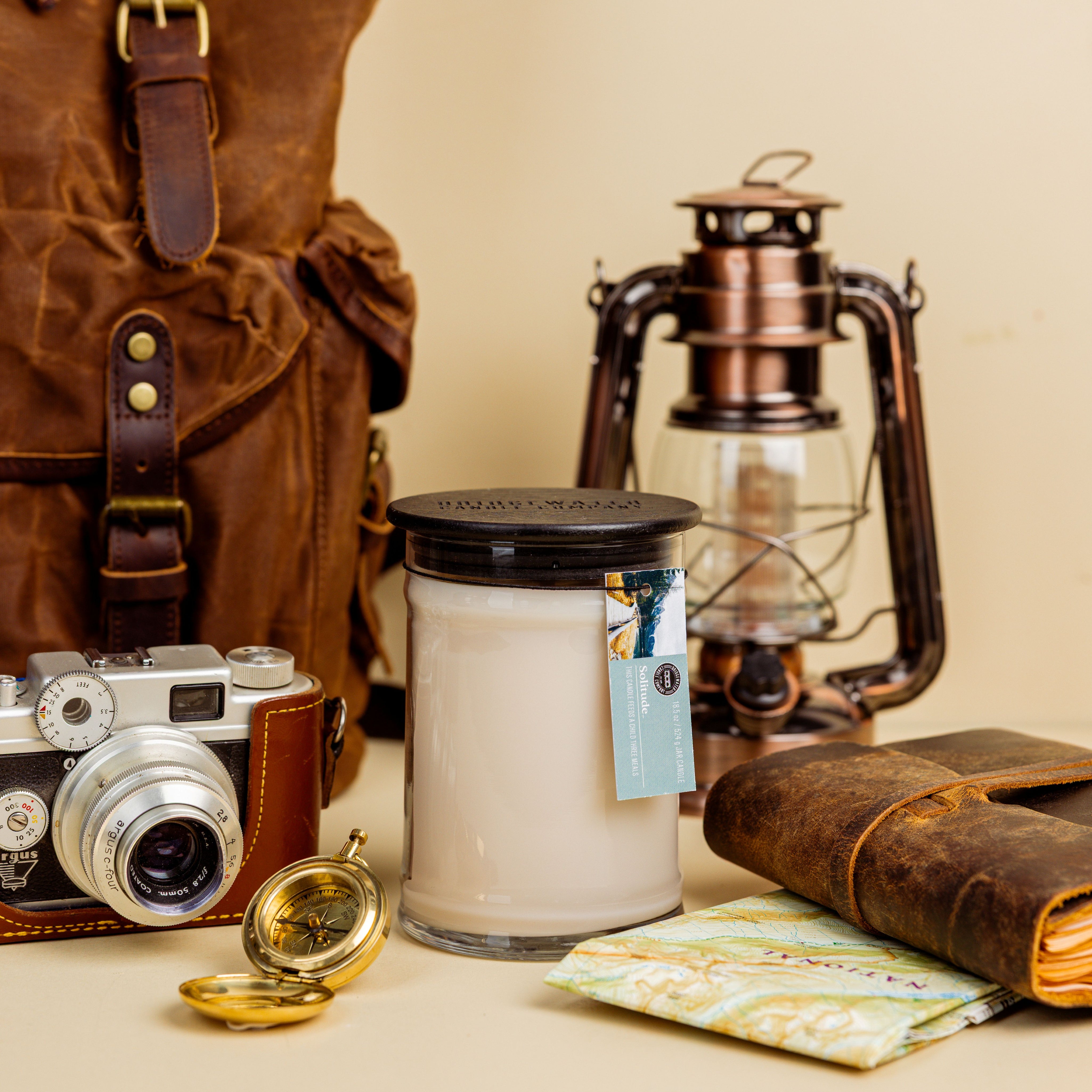A still life of travel items—vintage camera, brown backpack, compass, map, leather journal, Bridgewater Candle Solitude Large Jar Candle with soy-blend wax, and brass lantern—arranged on a beige background.