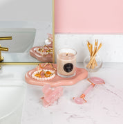 A bathroom counter features a pink tray with the Bridgewater Candle Sweet Grace Votive Candle, gold jewelry, a beaded bracelet, and a hair clip. Nearby are cotton swabs in a glass jar and a pink facial roller, creating a cozy vibe by the sink and mirror.