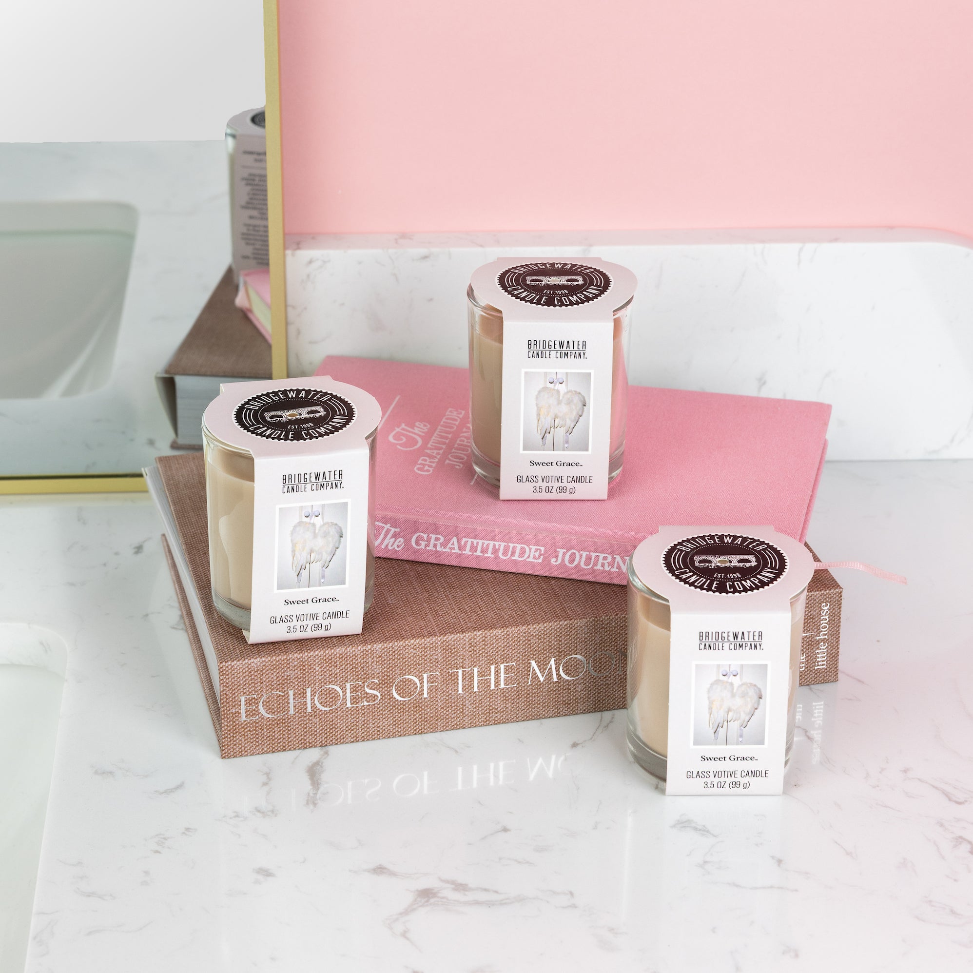 Three Bridgewater Candle Sweet Grace votive candles create a cozy bathroom ambiance—two rest on books, Echoes of the Moon and The Gratitude Journal, while one sits by a pink cloth—perfect for elevating your home fragrance.