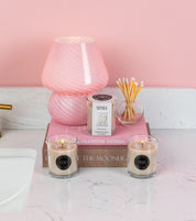A pink-striped glass lamp, a pink gratitude journal, Rise of the Moonlight, two Bridgewater Candle Sweet Grace votive candles, a boxed candle, and a round jar of cotton swabs create a cozy ambiance on the white bathroom counter.