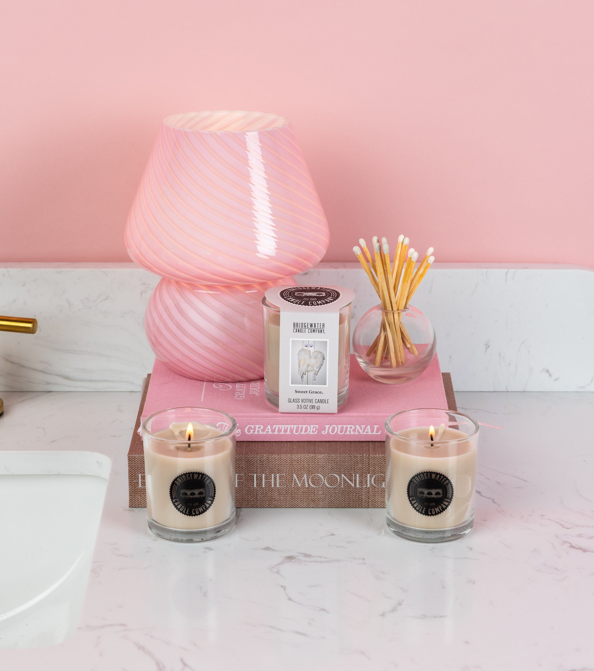 A pink-striped glass lamp, a pink gratitude journal, Rise of the Moonlight, two Bridgewater Candle Sweet Grace votive candles, a boxed candle, and a round jar of cotton swabs create a cozy ambiance on the white bathroom counter.