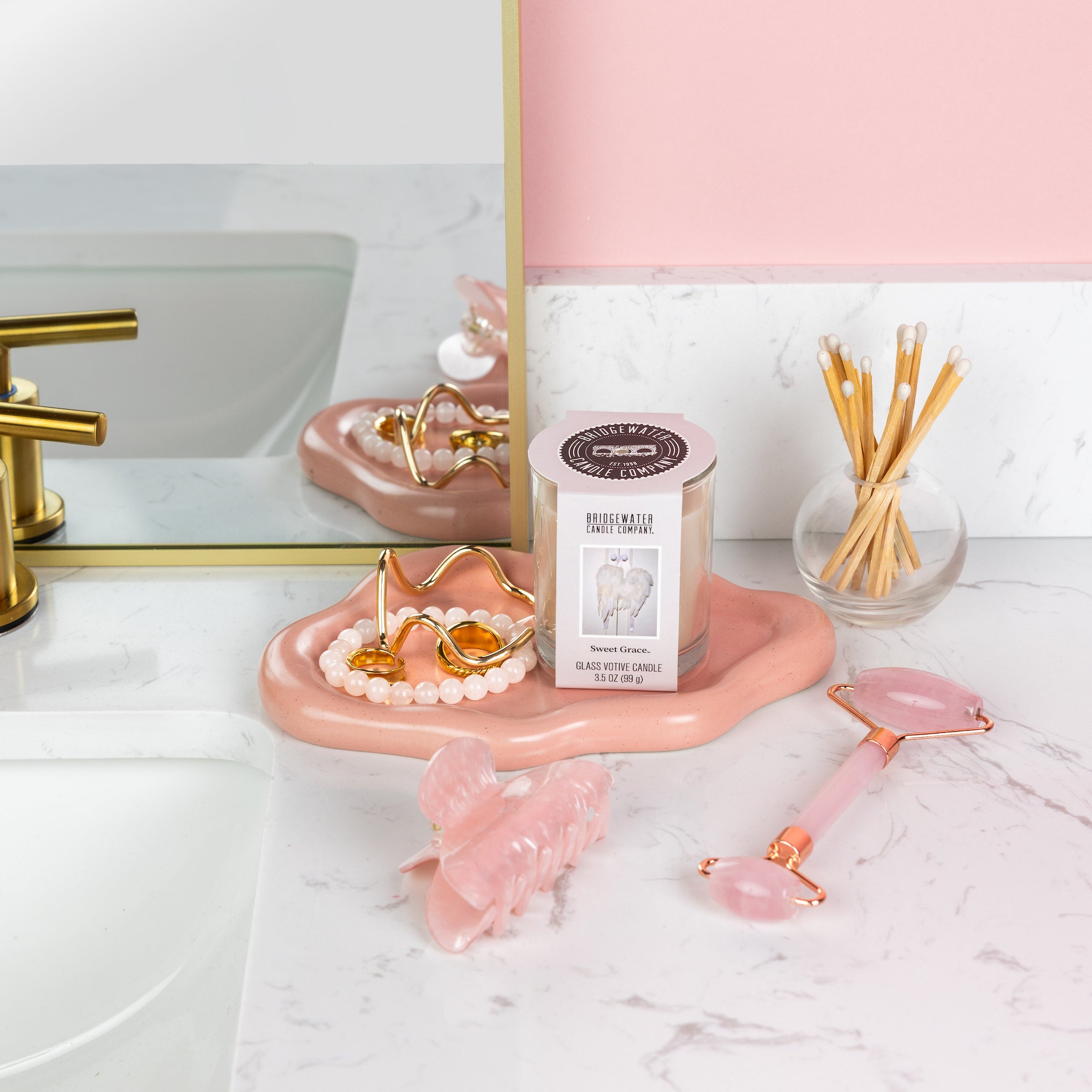 A pink and white bathroom countertop with a gold faucet, mirror, Bridgewater Candle's Sweet Grace Votive Candle for ambiance, pearl bracelet, gold jewelry, pink hair clip and facial roller, plus a glass jar of cotton swabs arranged neatly.
