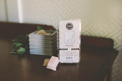 A package of scented wax bars labeled Sweet Grace sits on a wooden table next to a stack of small dishes and some greenery. A few wax pieces are placed in front of the package.