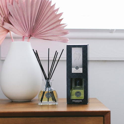 A reed diffuser and its box sit on a wooden table next to a white vase holding pink dried leaves, with a white window in the background.