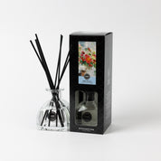 A glass High Bloom Reed Diffuser by Bridgewater Candle with black reeds stands next to its black box, decorated with a vibrant floral design and the label “High Room,” bringing premium fragrance and elegance. Plain white background.