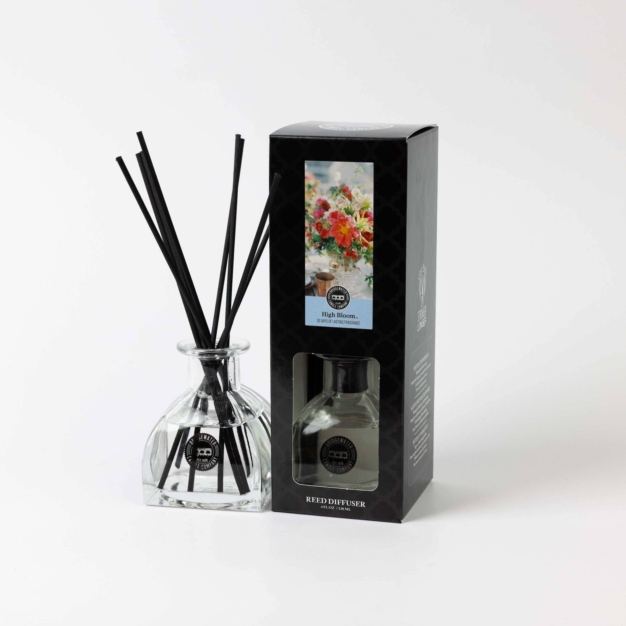 A glass High Bloom Reed Diffuser by Bridgewater Candle with black reeds stands next to its black box, decorated with a vibrant floral design and the label “High Room,” bringing premium fragrance and elegance. Plain white background.