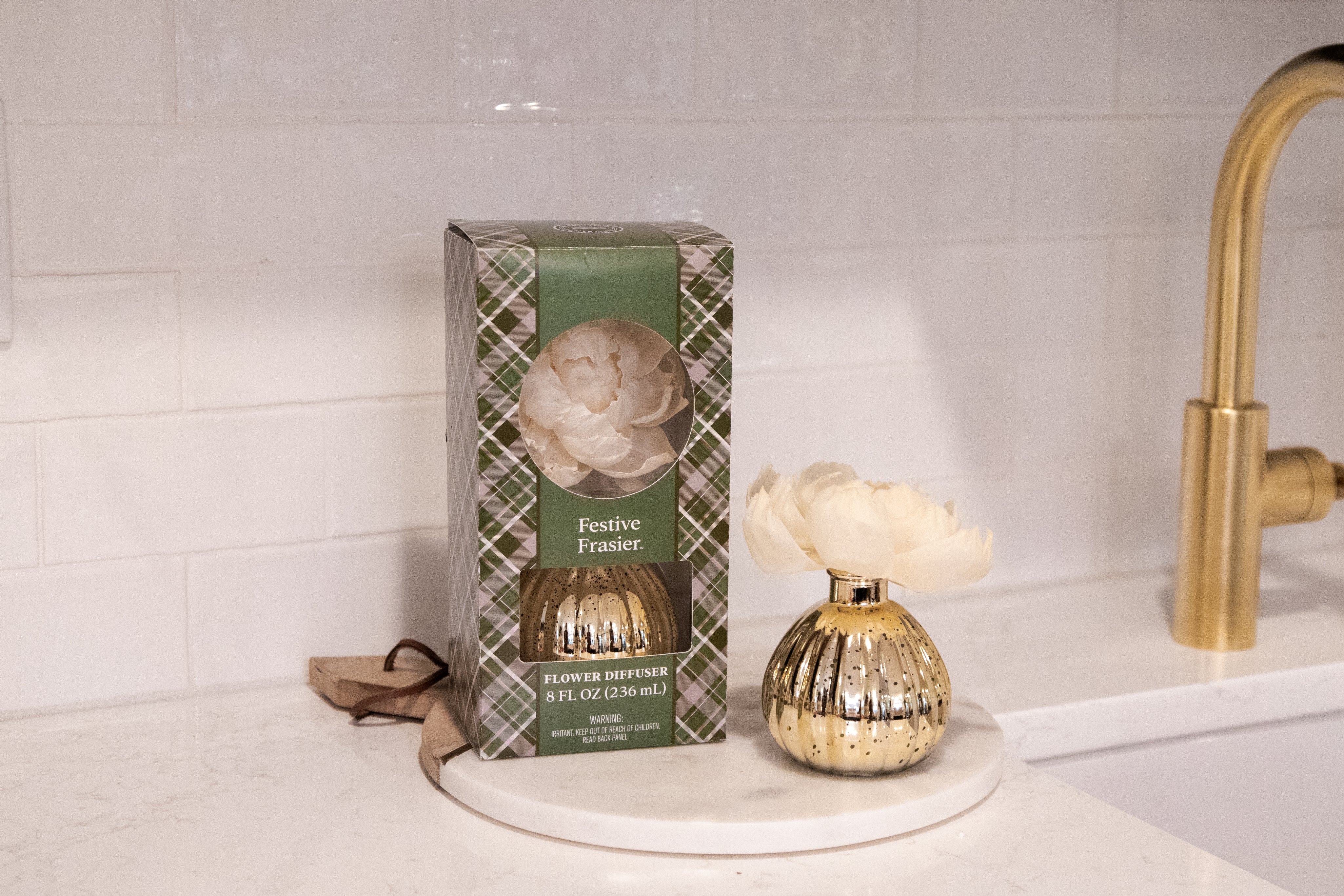 The Festive Frasier Holiday Mercury Flower Diffuser by Bridgewater Candle Company sits on a white kitchen countertop beside its green plaid box, adding elegant holiday decor near a gold faucet and white tiled backsplash.