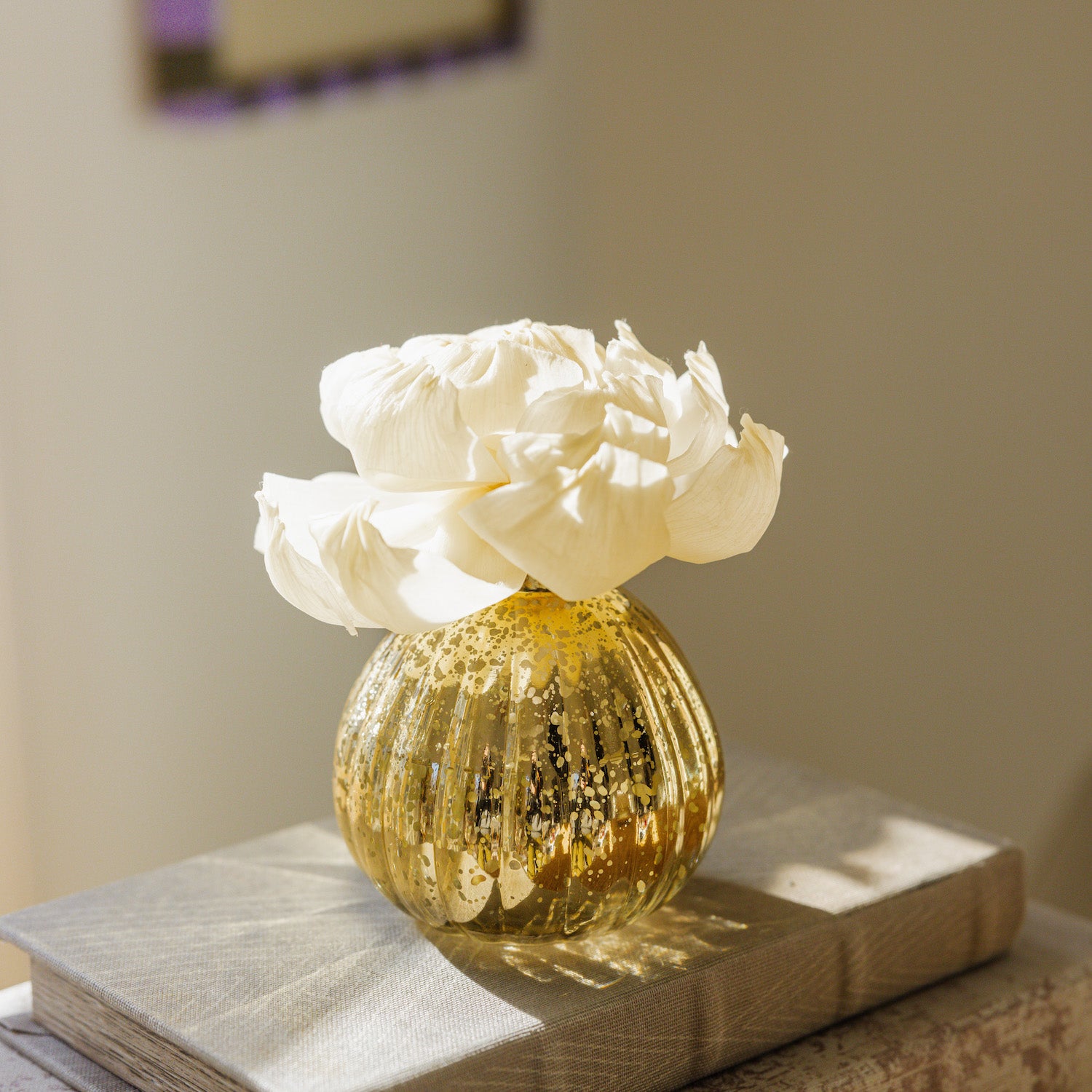 A small gold-speckled glass vase with white flowers and Bridgewater Candle’s Sweet Grace Dye-Free Flower Refill sits atop a textured book in gentle sunlight, infusing the serene scene with a long-lasting, dye-free fragrance.