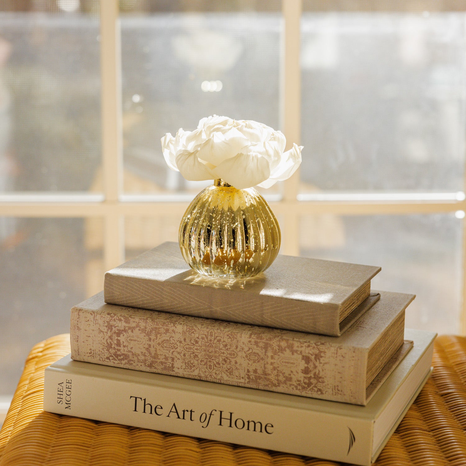 A small gold vase with white flowers and a Bridgewater Candle Sweet Grace Dye-Free Flower Refill for lasting fragrance rests atop three books, including The Art of Home, on a wicker surface by a sunlit window.