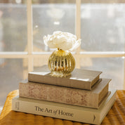 A small gold vase with white flowers and a Bridgewater Candle Sweet Grace Dye-Free Flower Refill for lasting fragrance rests atop three books, including The Art of Home, on a wicker surface by a sunlit window.
