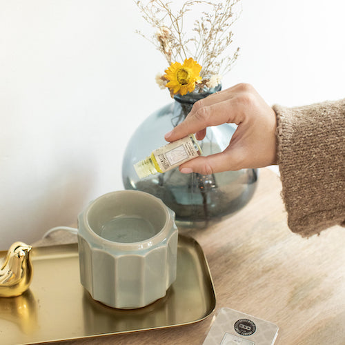 A hand pours essential oil into a ceramic diffuser on a gold tray. A blue vase with yellow flowers and a gold bird figurine are also on the wooden table.