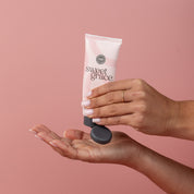 A person with manicured nails dispenses Bridgewater Candle Company Sweet Grace Hand Cream from a pink tube onto their palm, set against a solid pink background.