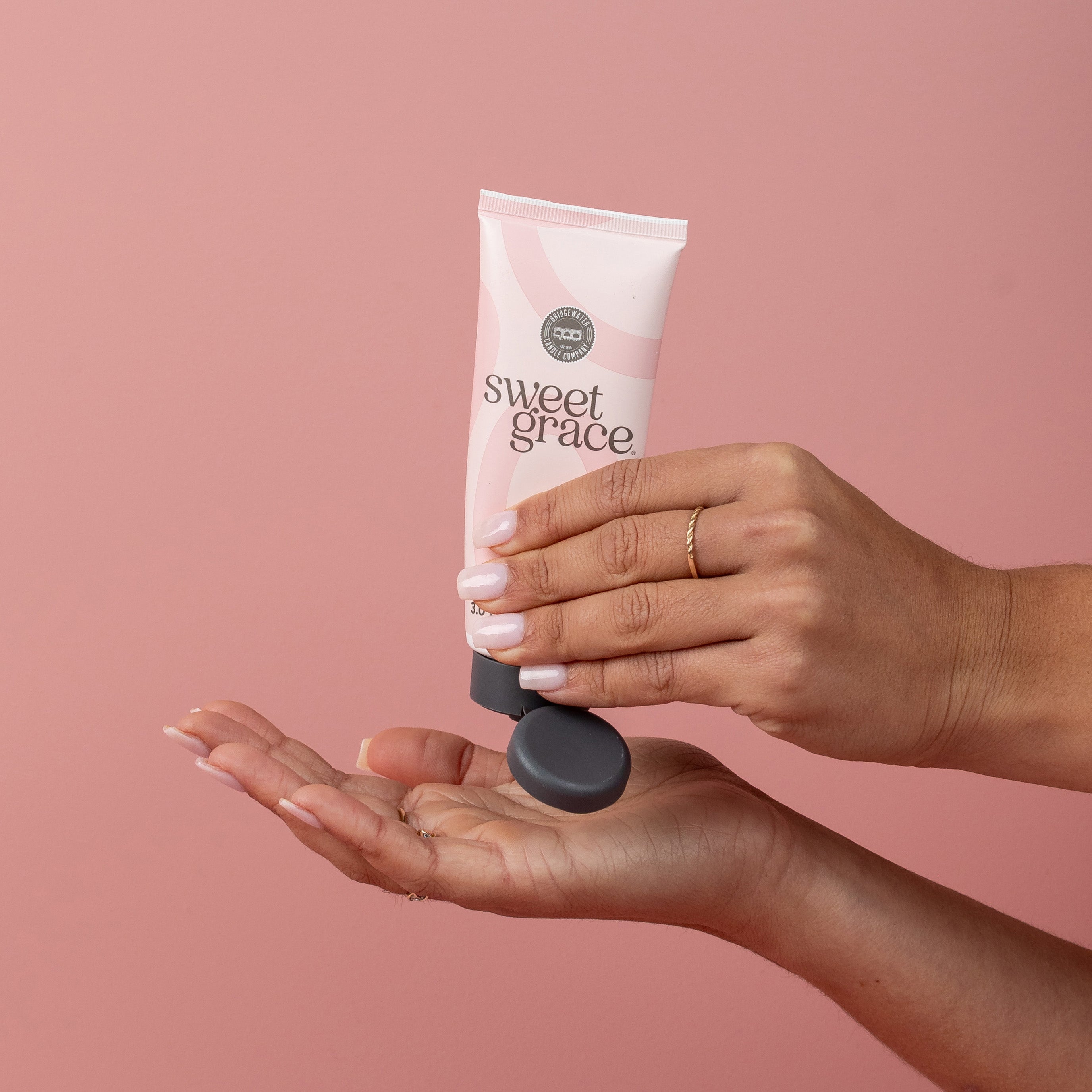 A person with manicured nails dispenses Bridgewater Candle Company Sweet Grace Hand Cream from a pink tube onto their palm, set against a solid pink background.