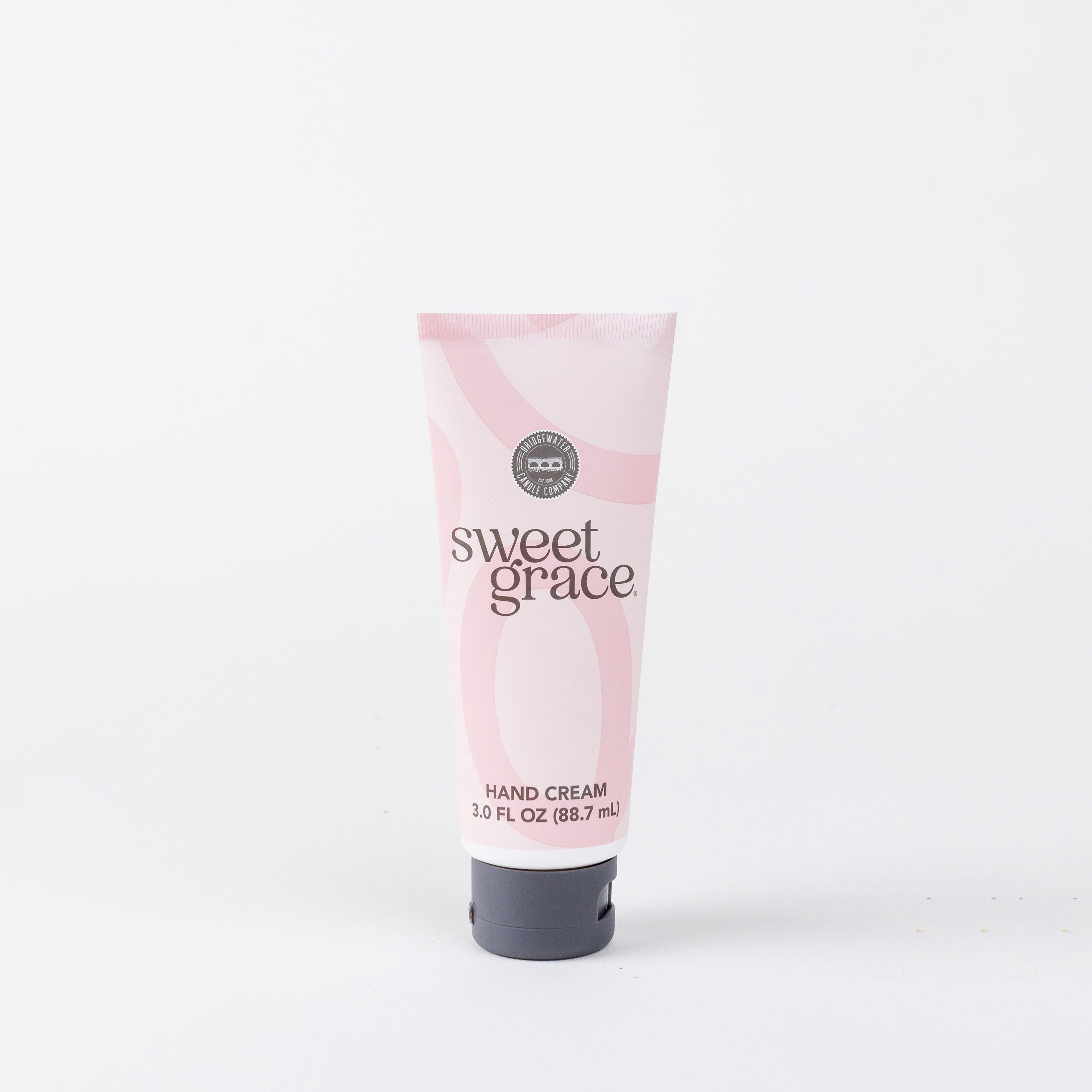 A 3.0 fl oz (88.7 mL) pink tube of Bridgewater Candle Company’s Sweet Grace Hand Cream stands upright against a white background, displaying black and white text with a swirl design that highlights its fragrant, elegant appeal.