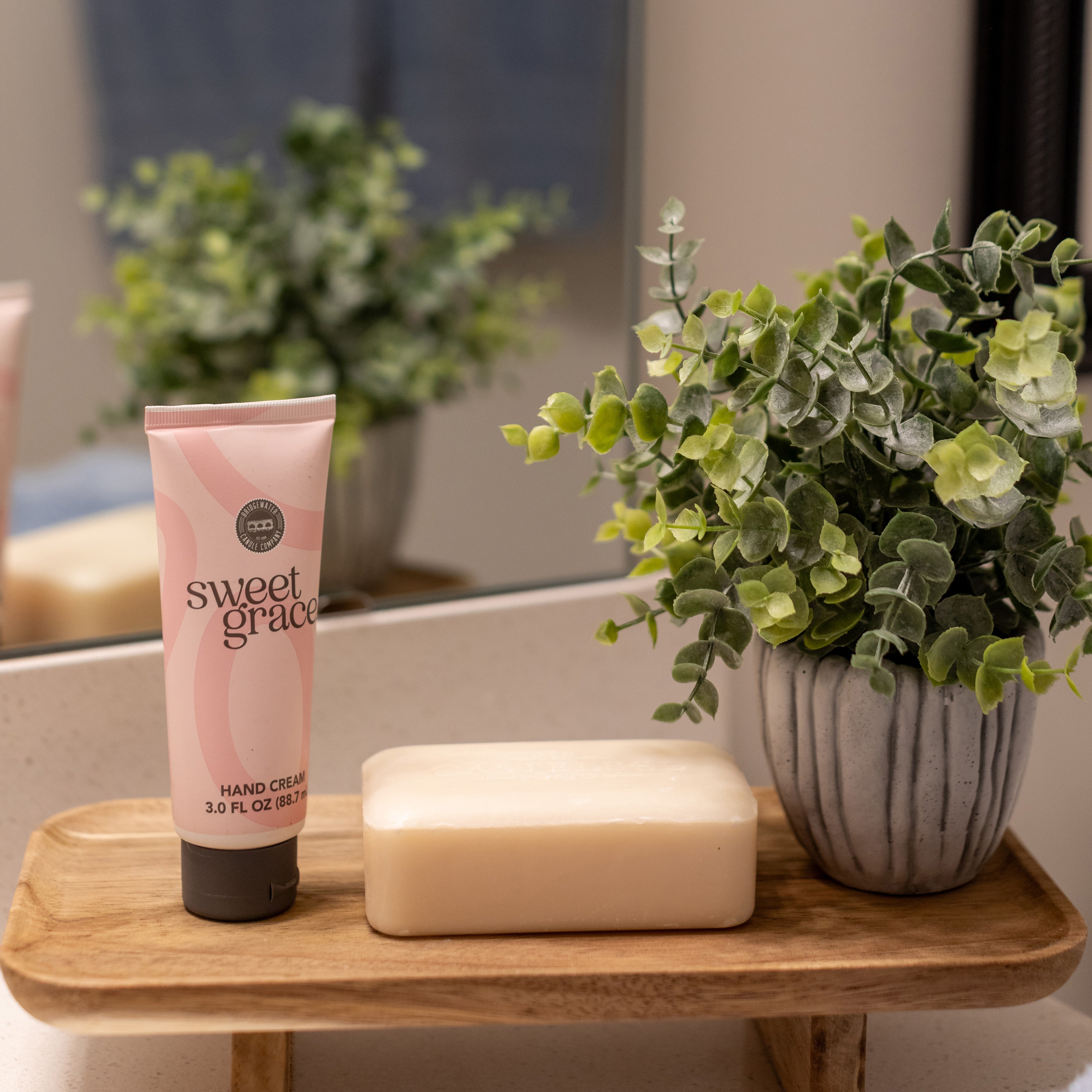 A wooden tray on a bathroom counter holds a tube of Sweet Grace Hand Cream by Bridgewater Candle Company, a bar of soap, and a small potted artificial plant. A mirror and blurred greenery appear in the background.