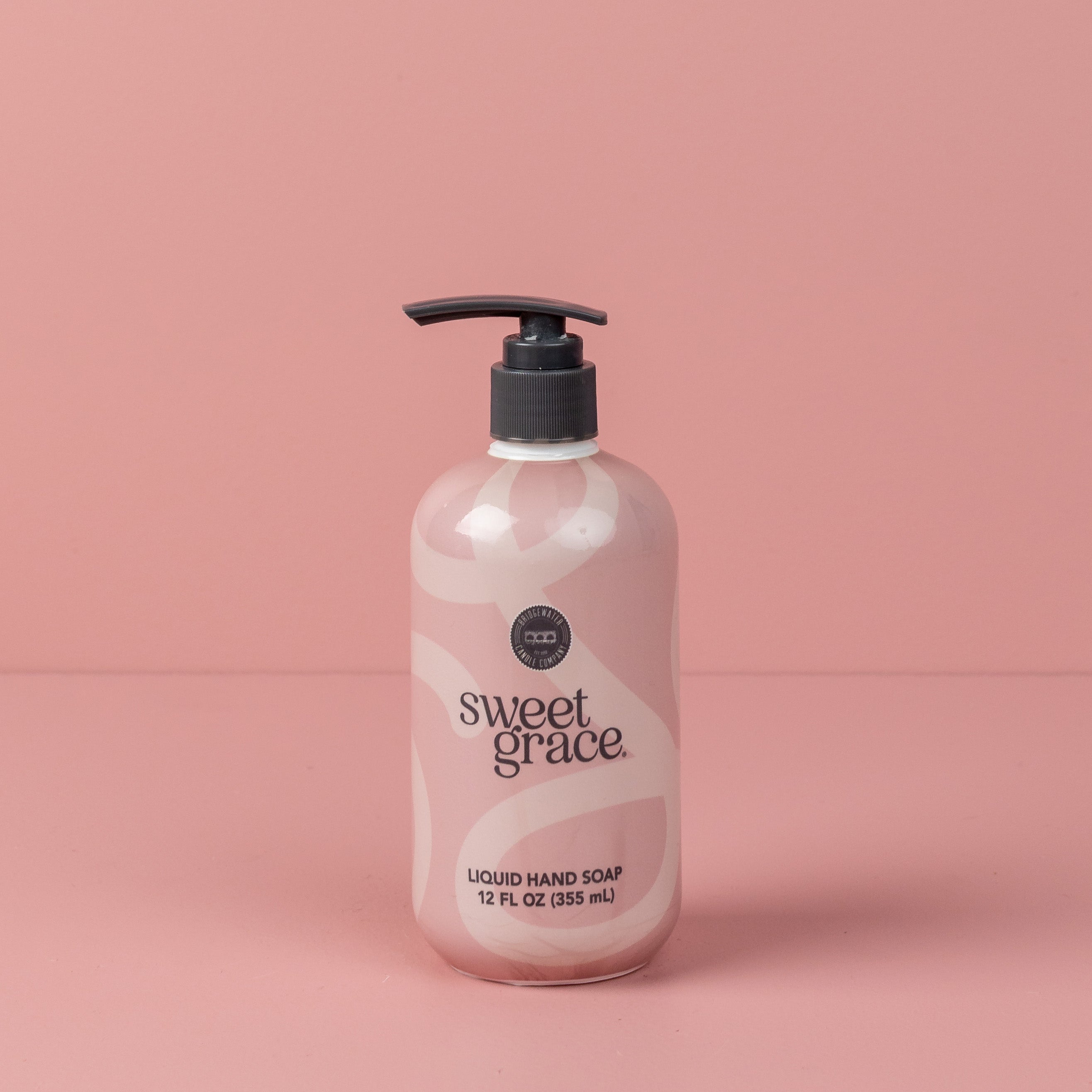 A bottle of Bridgewater Candle Companys Sweet Grace Liquid Soap with a pump dispenser and a pink and white abstract design sits against a matching pink background.