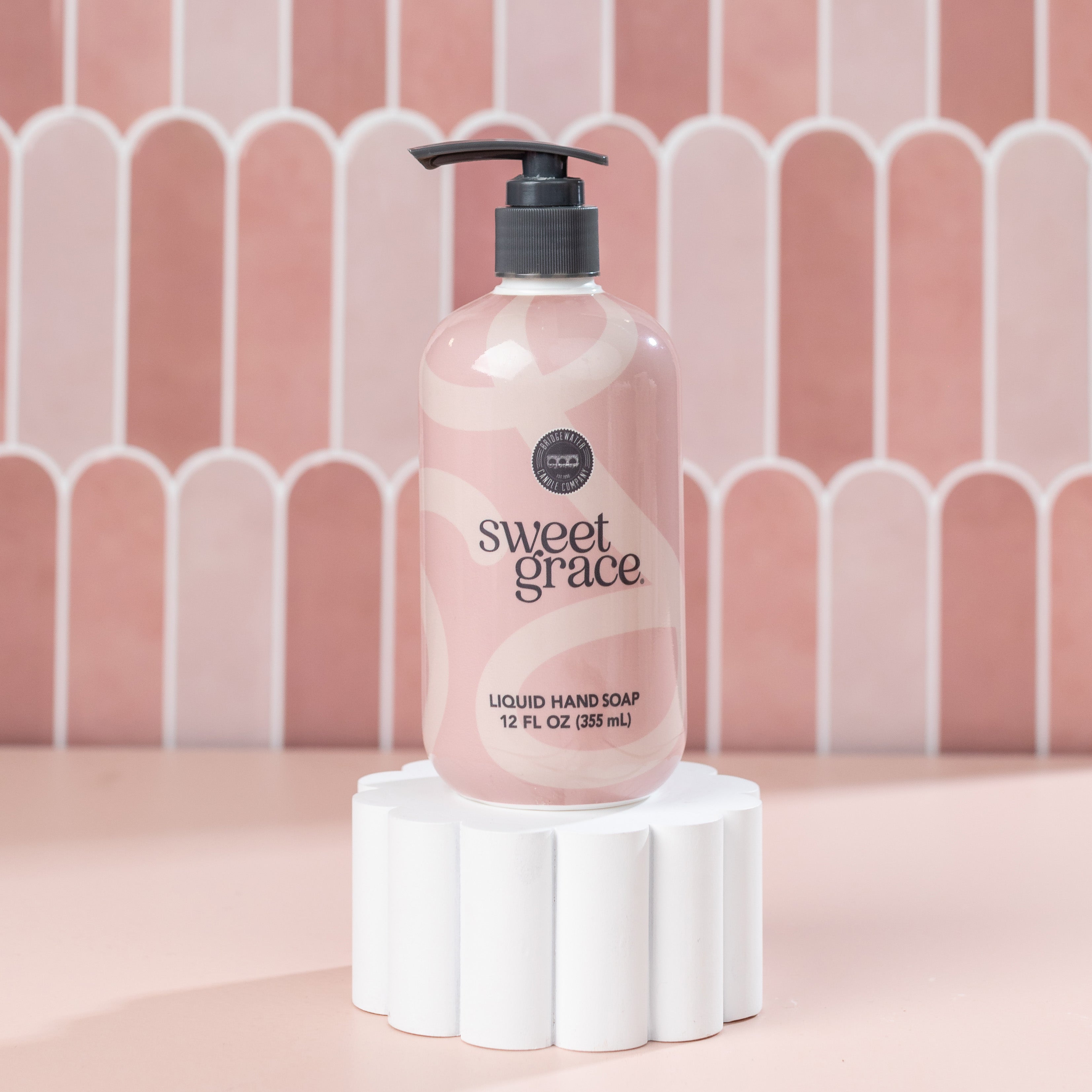 A bottle of Bridgewater Candle Companys Sweet Grace Liquid Soap with a black pump rests on a white pedestal, set against pink, scalloped tiles. The light pink bottle features abstract white lines and offers a fragrant handwashing experience.