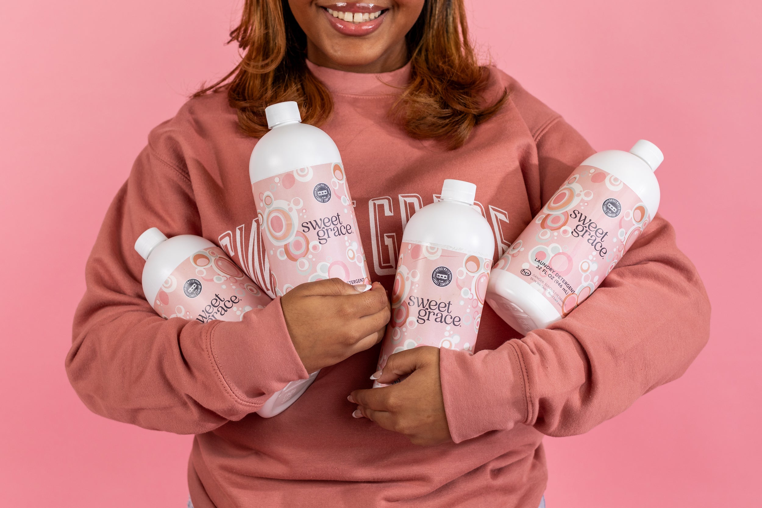 A person in a pink sweatshirt smiles while holding four bottles labeled Sweet Grace against a pink background.