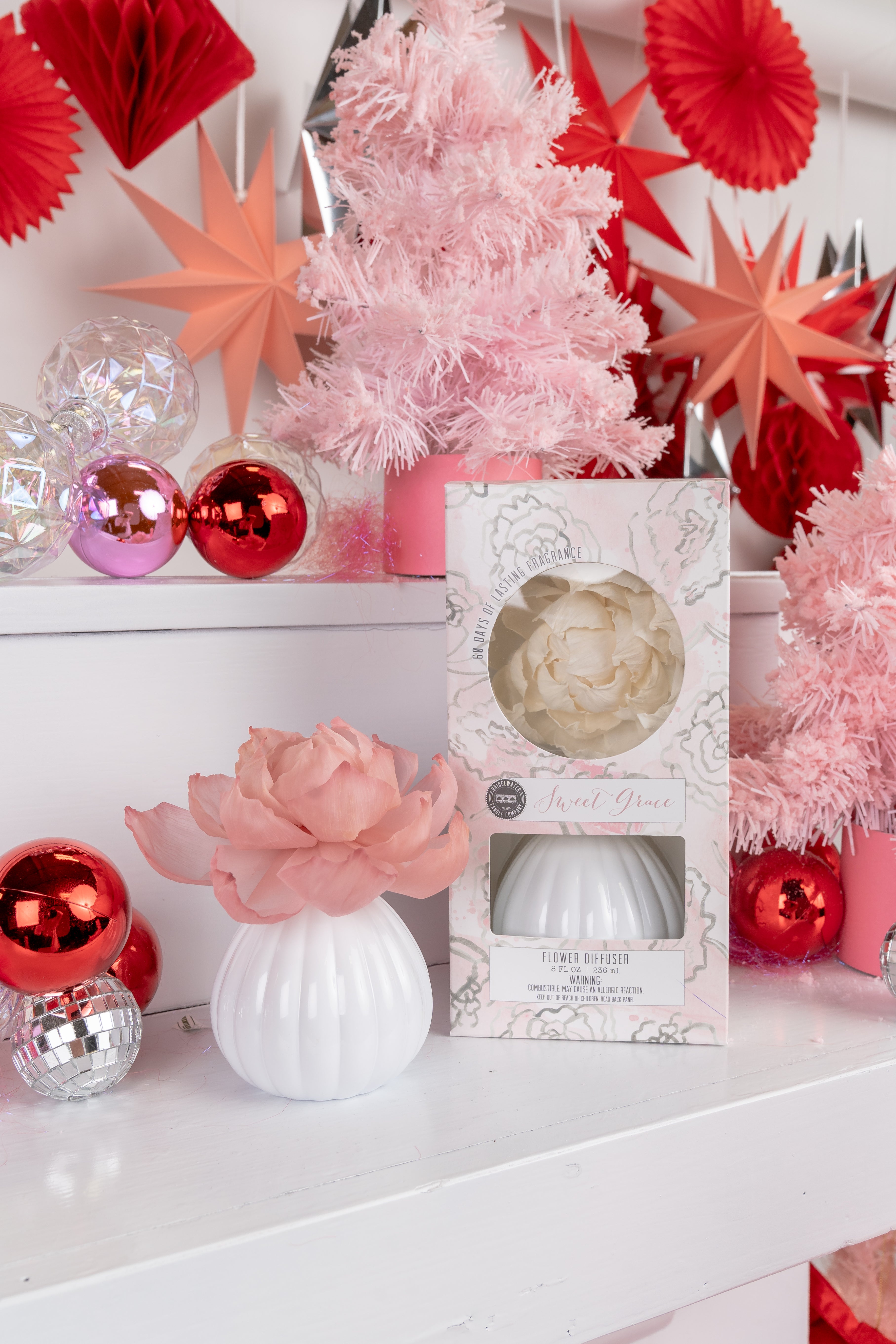 A white flower diffuser with a pink flower sits on a white shelf next to its floral-patterned box. The background features pink trees, red ornaments, silver stars, and pink paper decorations.