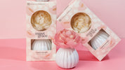 Three white ceramic Sweet Grace Flower Diffusers by Bridgewater Candle Company, each with a pink artificial flower, are displayed on a pink surface. Two sit in pastel pink-patterned boxes; one is unboxed in front, showcasing its elegant beauty and scent.