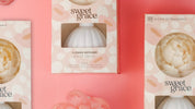 Gift boxes of the Bridgewater Candle Company Sweet Grace Flower Diffuser are displayed on a pink backdrop, encircled by artificial pink and white flowers. Each pastel box has a round cutout showing the diffuser and suggesting its beautiful scent.