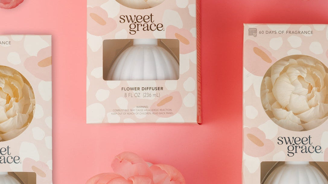 Gift boxes of the Bridgewater Candle Company Sweet Grace Flower Diffuser are displayed on a pink backdrop, encircled by artificial pink and white flowers. Each pastel box has a round cutout showing the diffuser and suggesting its beautiful scent.