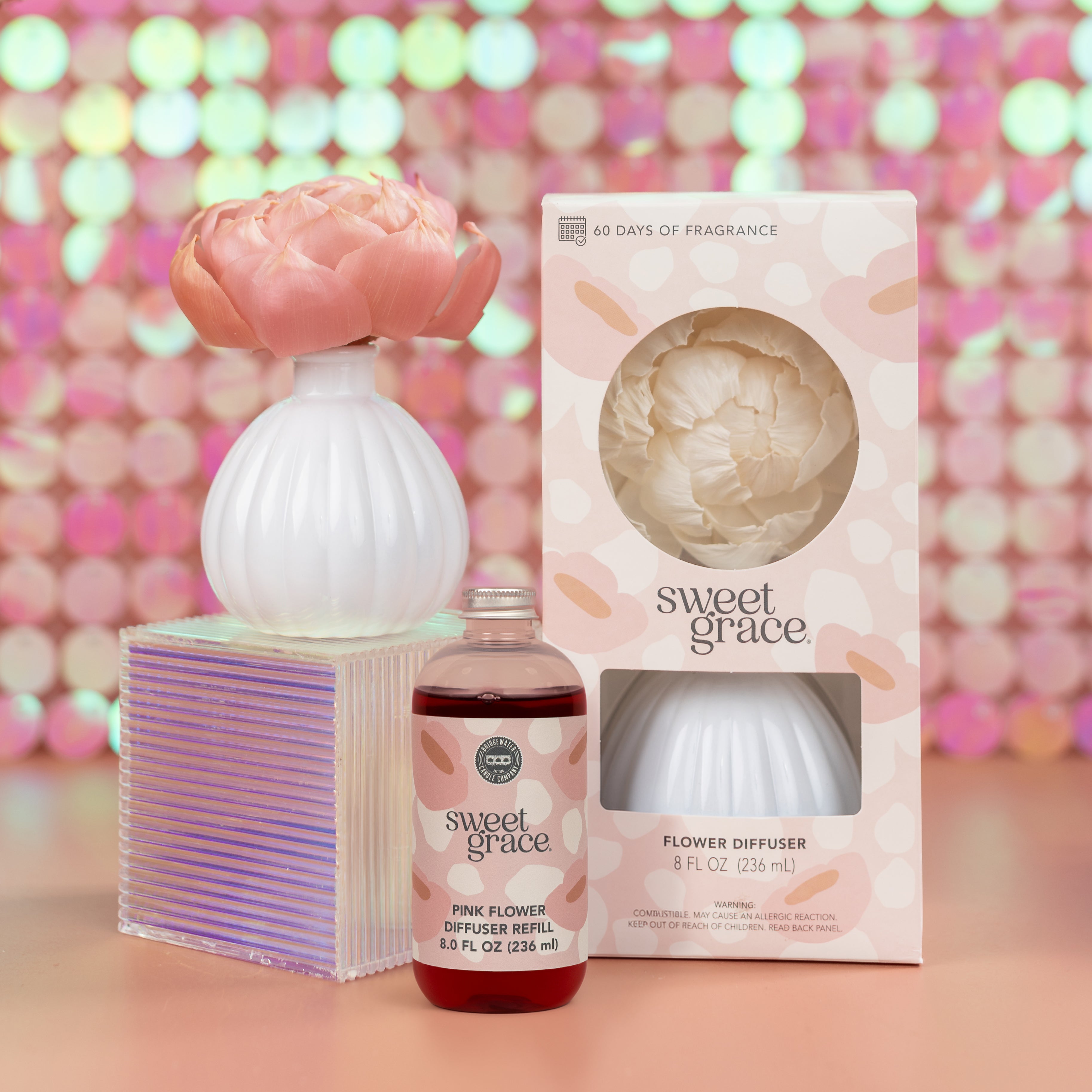 The Fast Bundle Sweet Grace Flower Diffuser & Refill Bundle, with product packaging, is displayed on a reflective surface against a pink bokeh background—ideal for a luxurious fragrance gift.