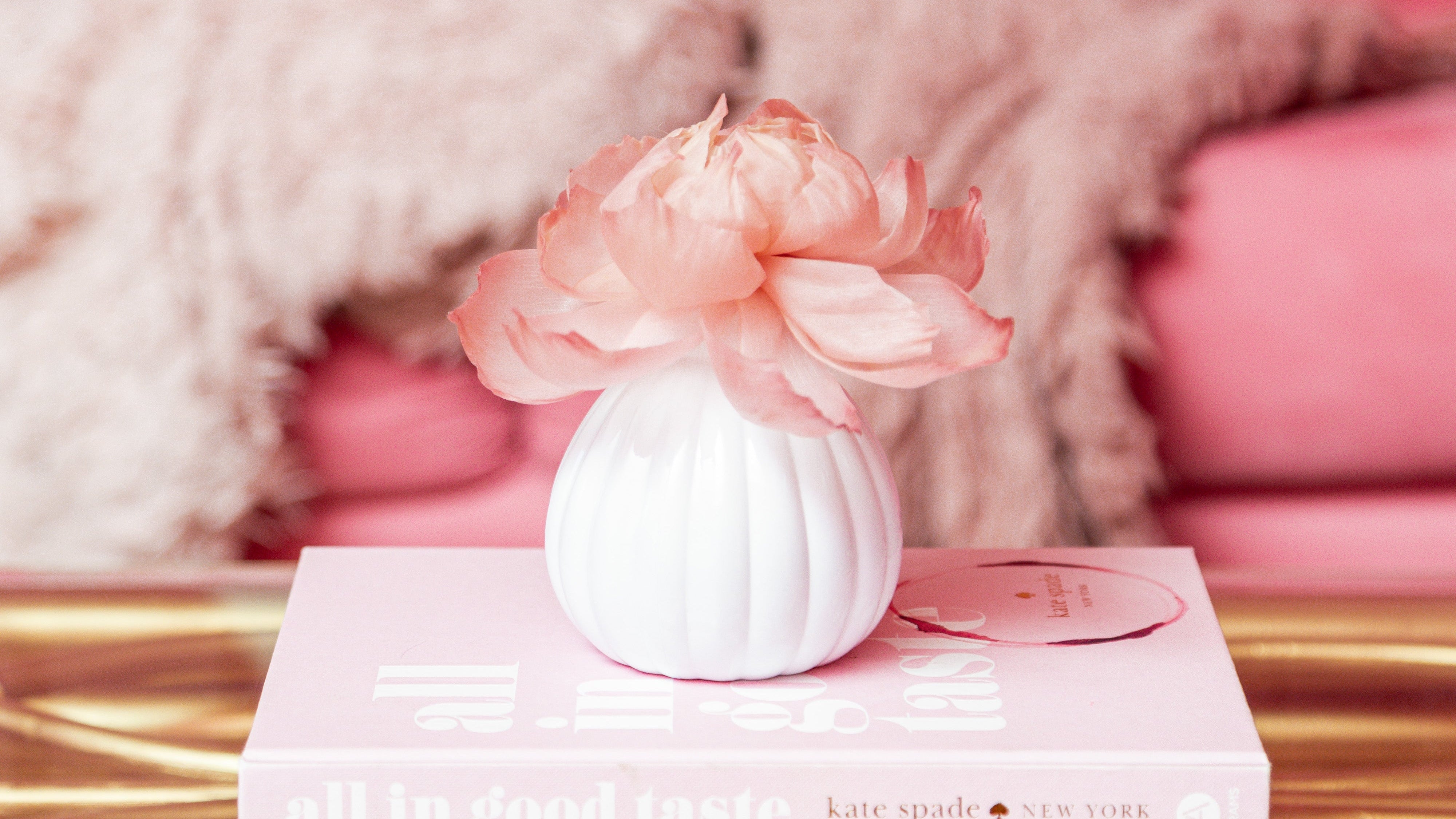 The Bridgewater Candle Company Sweet Grace Flower Diffuser in a white vase with a pink peony sits on stacked books atop a gold table, with a pink sofa and soft blanket behind it, infusing the space with its inviting fragrance.
