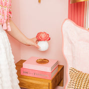 A hand places a Bridgewater Candle Company Sweet Grace Flower Diffuser with a pink bloom on a wooden nightstand beside two stacked pink books in a pink-themed room featuring a pink pillow and gold accents.
