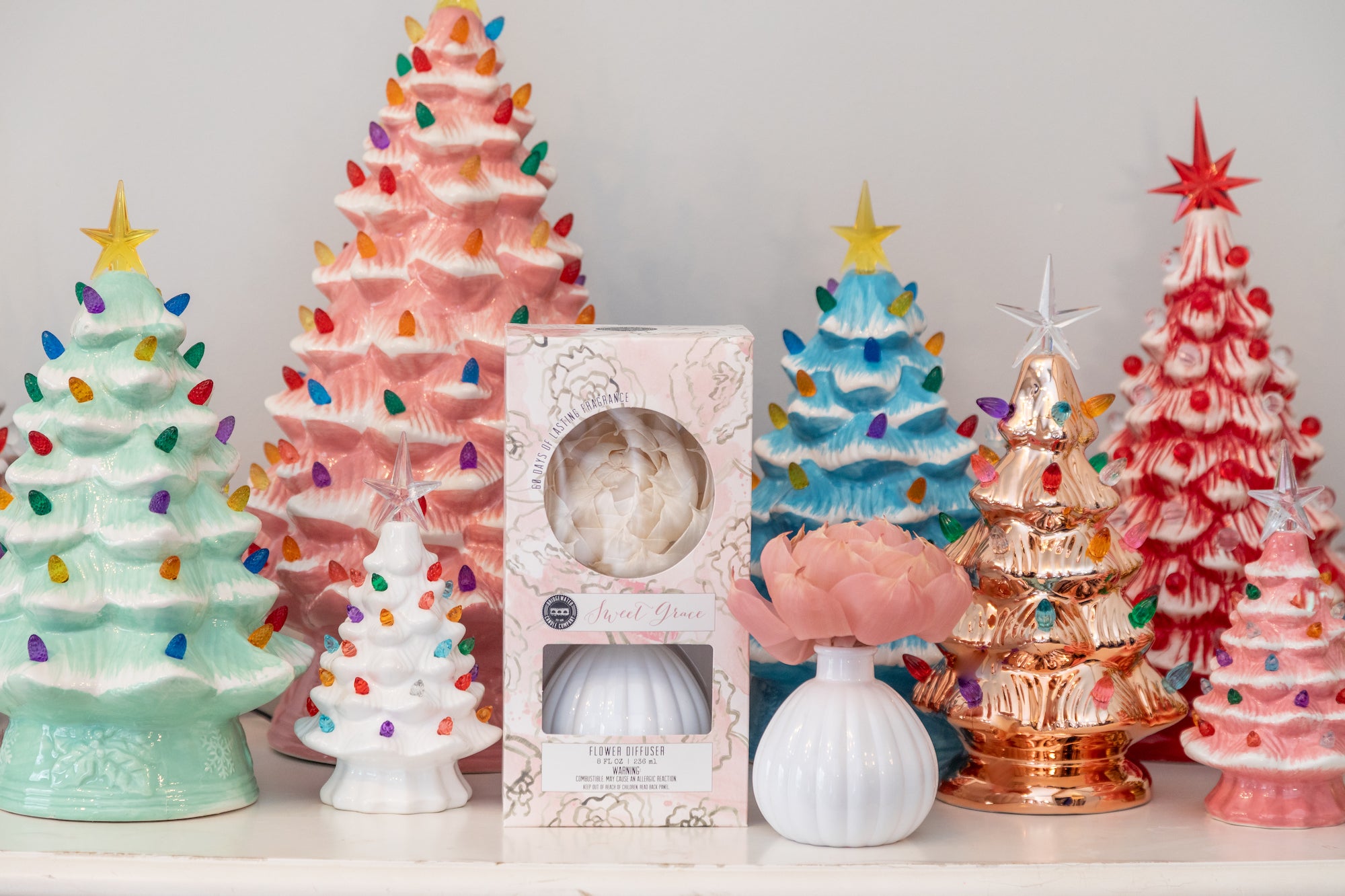A display of ceramic Christmas trees in pastel colors, some with multicolored lights and stars on top, with a boxed flower diffuser and a small white vase holding a pink flower in the foreground.
