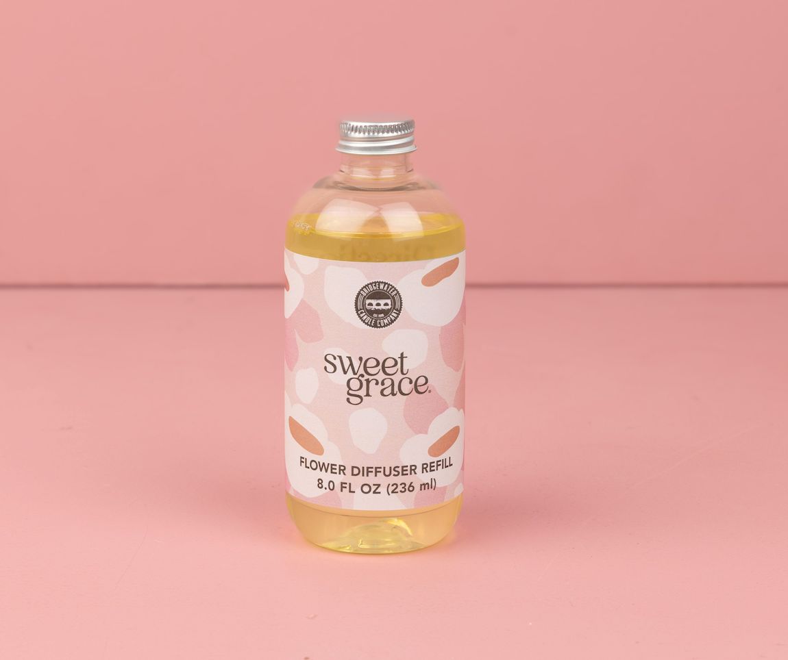 A clear bottle of Bridgewater Candles Sweet Grace Dye-Free Flower Refill, filled with dye-free yellow liquid, silver cap, and a pink-white label, sits on a pink surface and delivers long-lasting fragrance against a pink background.