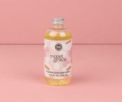 A clear bottle of Bridgewater Candles Sweet Grace Dye-Free Flower Refill, filled with dye-free yellow liquid, silver cap, and a pink-white label, sits on a pink surface and delivers long-lasting fragrance against a pink background.