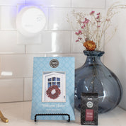 A blue sachet labeled Welcome Home and the Bridgewater Candle Companys Pura+Bridgewater Fragrance Refill are displayed with a white smart diffuser in front of a blue vase with dried flowers, all set against a tiled wall.